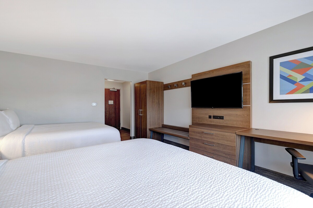 Habitación de hotel con dos camas, un escritorio de madera, un televisor de pantalla plana y un cuadro abstracto enmarcado en la pared.