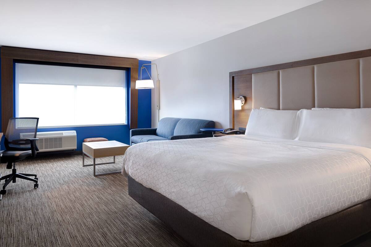 Une chambre d’hôtel avec un grand lit, un bureau avec un ordinateur, une chaise et une fenêtre avec des stores.