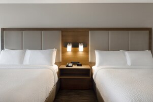 Una habitación de hotel con dos camas, un cabecero de madera, una mesita de noche con teléfono y reloj, y una lámpara de pared.