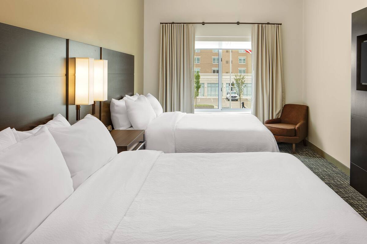Ein Hotelzimmer mit zwei Betten, einem Fernseher, einem Sessel und einem Fenster mit Vorhängen.