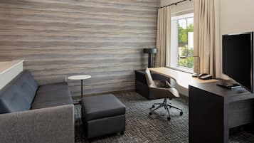 A modern hotel room with a grey sofa, a small round table, a desk with a chair, a TV, and a window with curtains.