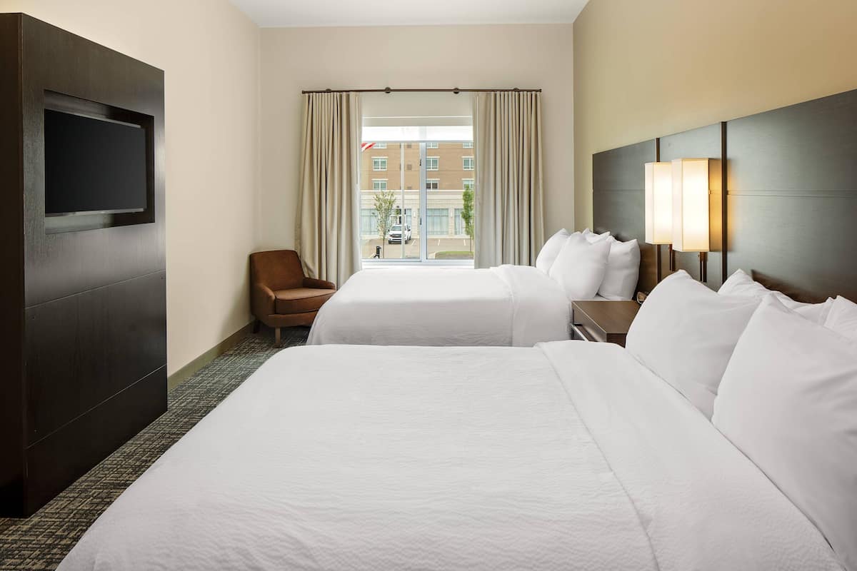 Ein Hotelzimmer mit zwei Betten, einem Fernseher, einem Sessel und einem Fenster mit Vorhängen.