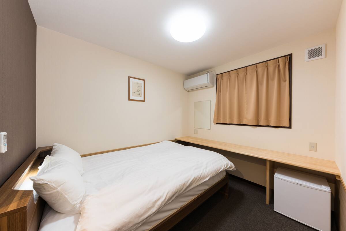 Una cama individual con estructura de madera, una mesita de noche pequeña, un aire acondicionado instalado en la pared, una ventana con cortinas color beige y un cuadro enmarcado en la pared.