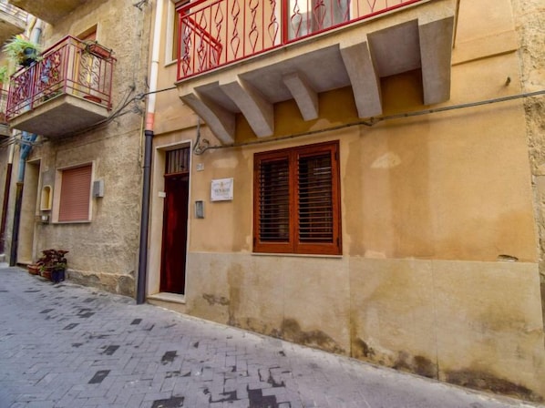 Une rue étroite bordée d’un immeuble doté d’un balcon orné de balustrades en métal ouvragé et d’un volet de fenêtre en bois.