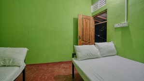 Une pièce aux murs verts, avec deux lits simples dotés de draps blancs, une porte en bois et une fenêtre avec une grille d’aération.