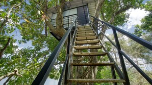 Un escalier en bois mène à une cabane perchée dans un arbre, et une rambarde en métal l’entoure, le tout noyé dans un feuillage verdoyant et luxuriant.