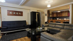Una sala de estar con un sofá negro y un sofá gris, una mesa de centro de cristal, y una zona de cocina con armarios de madera y electrodomésticos modernos.