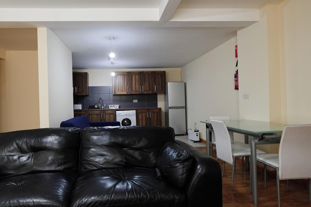 A living room with a black leather sofa, a dining table with white chairs, and a kitchen in the background.