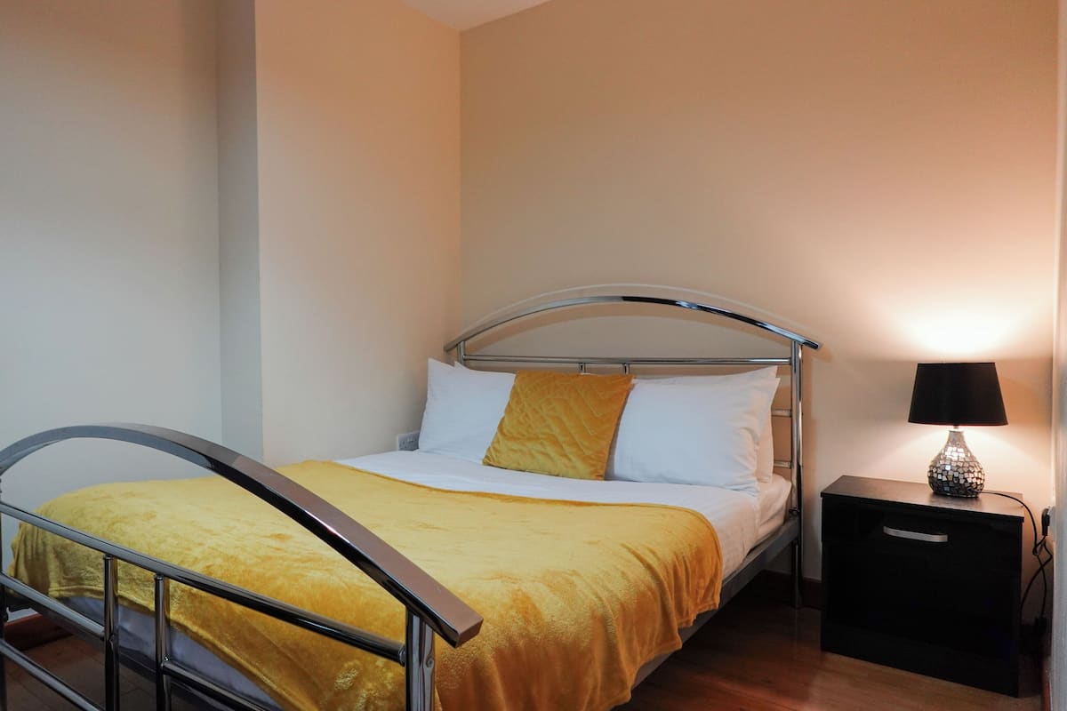 A single bed with a metal frame and a yellow bedspread, a bedside table with a lamp, and a plain wall.