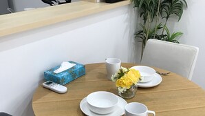 A round wooden dining table with white plates, a white mug, a spoon, and a fork. There is a blue box with a floral design and a small vase with yellow flowers.