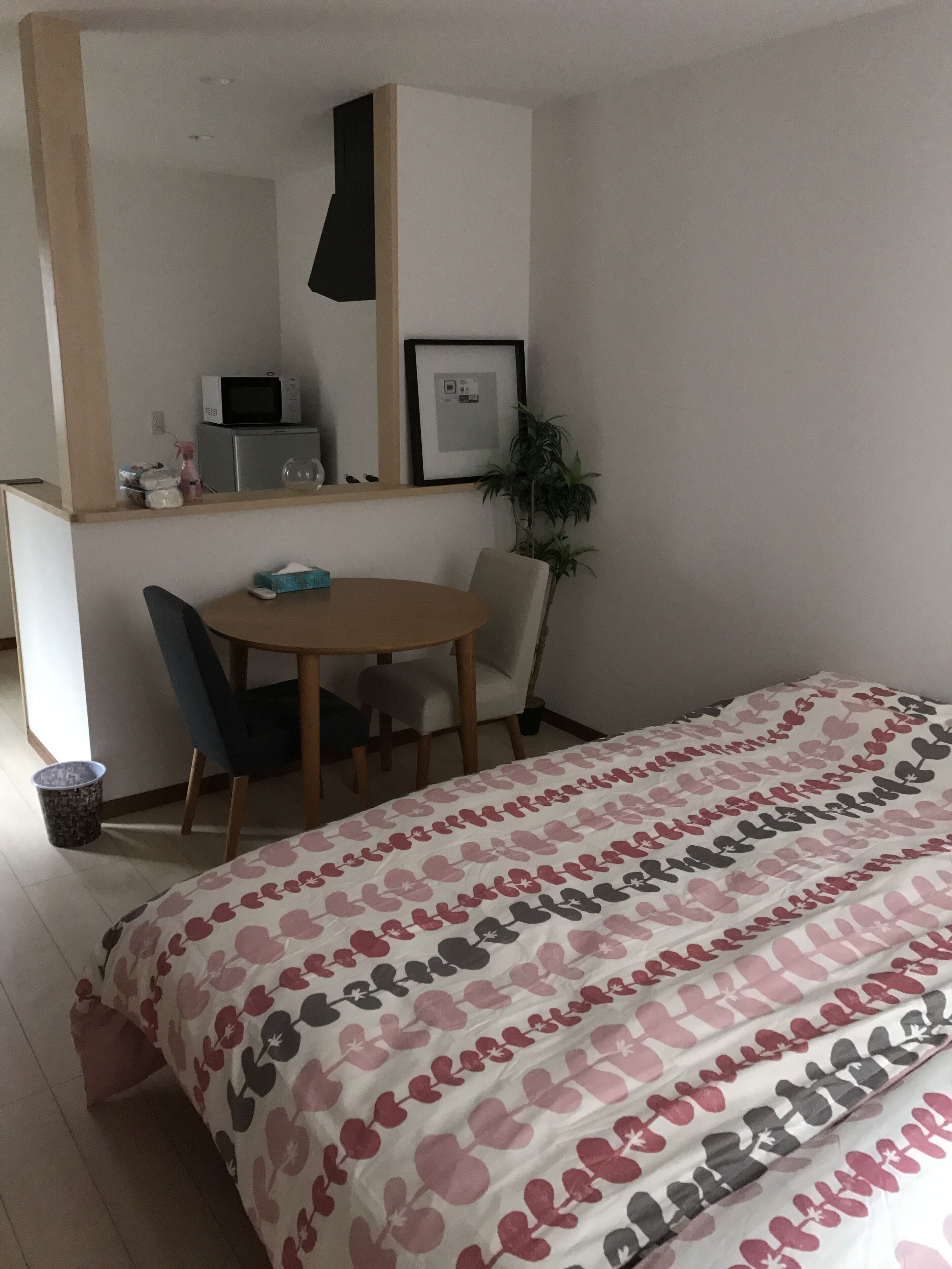 A bedroom with a bed, a desk, a chair, a microwave, a framed picture, and a potted plant.