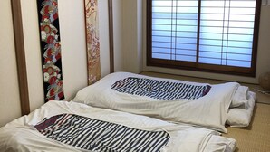 Una habitación japonesa tradicional con suelo de tatami, dos camas con ropa de cama a rayas, una ventana con celosías japonesas y rollos decorativos colgados.