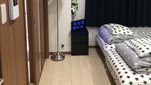 A hotel room with a bed, a bedside table with a digital clock, a floor lamp, a wall-mounted thermostat, and a window with dark curtains.