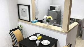 A compact dining area with a black table, two chairs, and a small side table with a vase of yellow flowers. A microwave and a refrigerator are visible in the background.