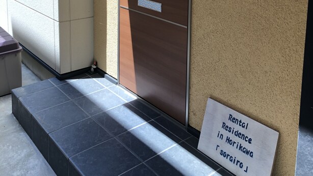 A rental residence entrance with a wooden door, a security camera, and a sign indicating the location in Horikawa, Sogairo.