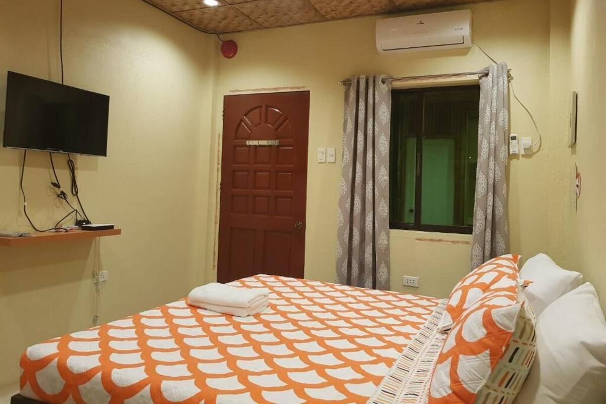 Un dormitorio con cama, televisor, equipo de aire acondicionado y ventana con cortinas.