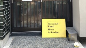 A traditional rental house entrance with a metal gate, a yellow signboard, and a small table with a cushion. - Kanazawa-Hachitabi Sennichi (Kanazawa)