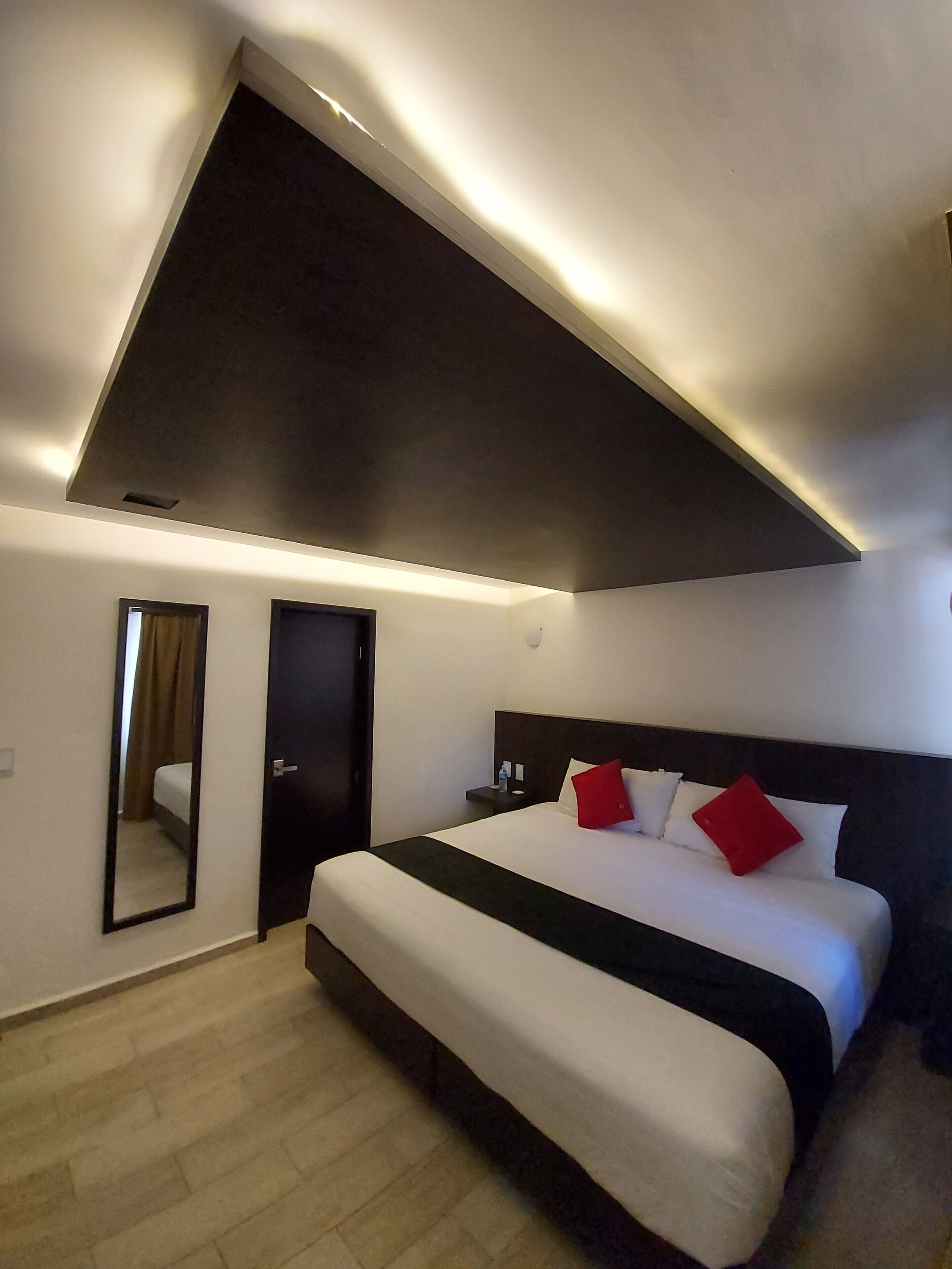 A modern hotel room with a large bed, a dark ceiling, a small bedside table with a lamp, and a mirror on the wall.