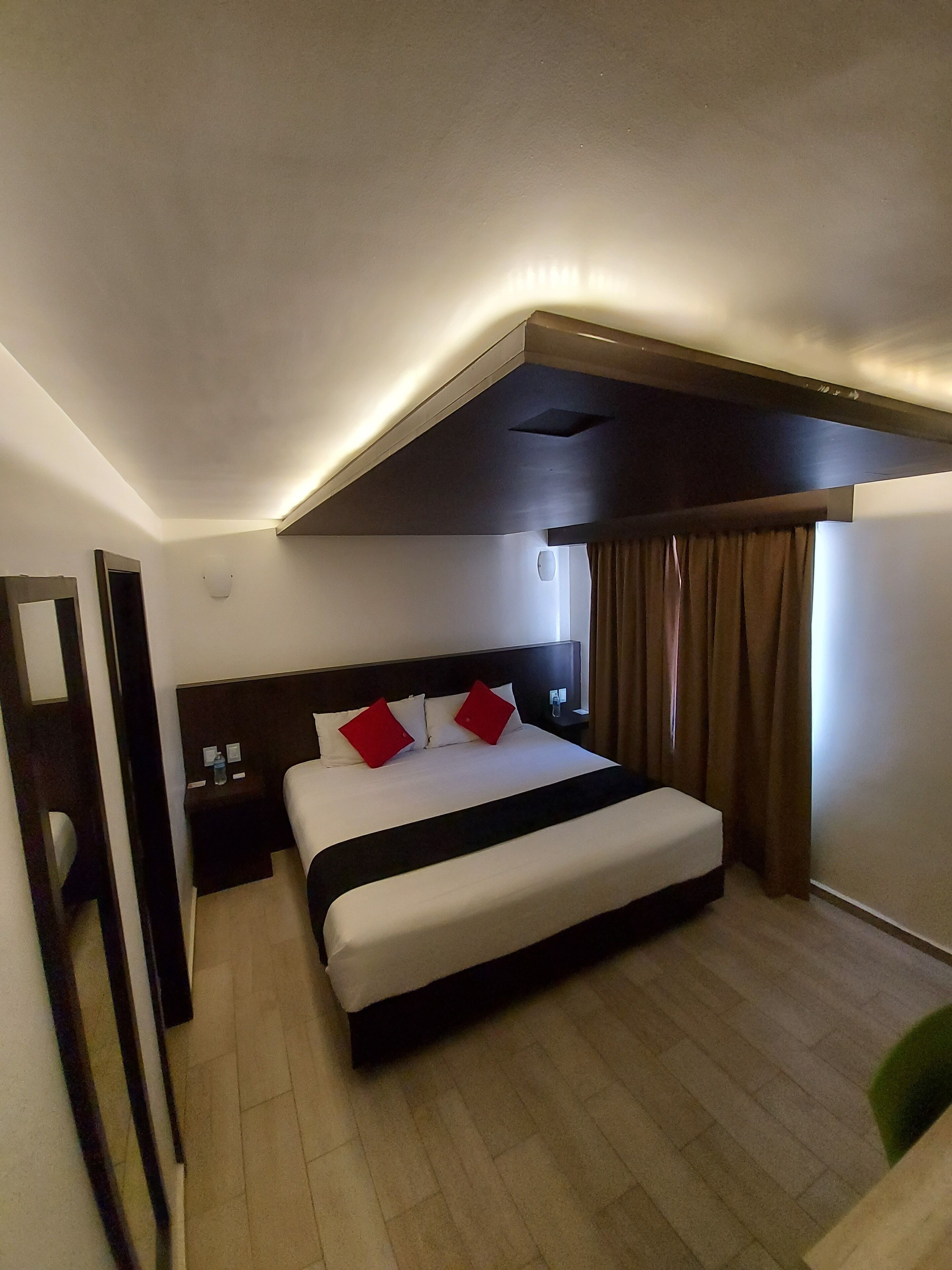 Una habitación de hotel con una cama grande, una mesita de noche, un espejo y una ventana con cortinas.