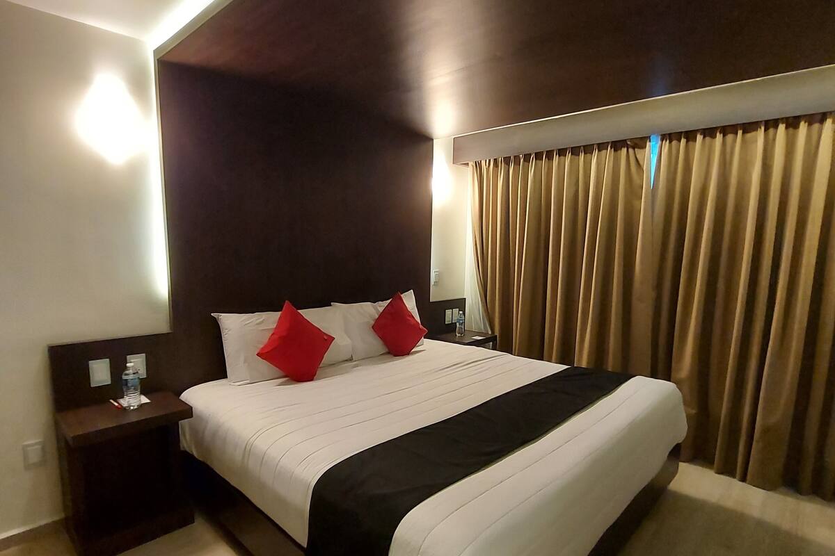 Een hotelkamer met een groot bed, nachtkastje, een wandlamp en een raam met gordijnen.