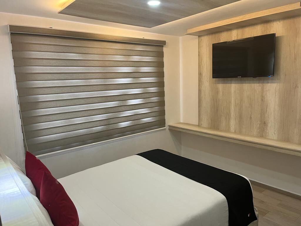 Un dormitorio con cama, televisor montado en la pared y ventana con persianas.