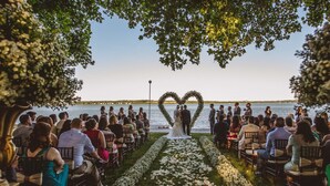 Una ceremonia de boda con invitados sentados en sillas dispuestas en filas, frente a un pasillo floral en forma de corazón que conduce a una pareja bajo un árbol.