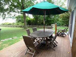 Una terraza de madera con una mesa y sillas, un parasol verde y un muro de piedra al fondo.