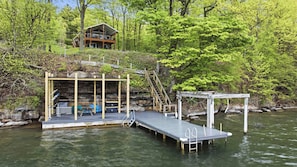 A wooden dock extending into a river, a small wooden structure with a table and benches, and a house on a hill in the background.