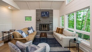 A cozy living room with a stone fireplace, two sofas, a coffee table, and large windows offering a view of trees.