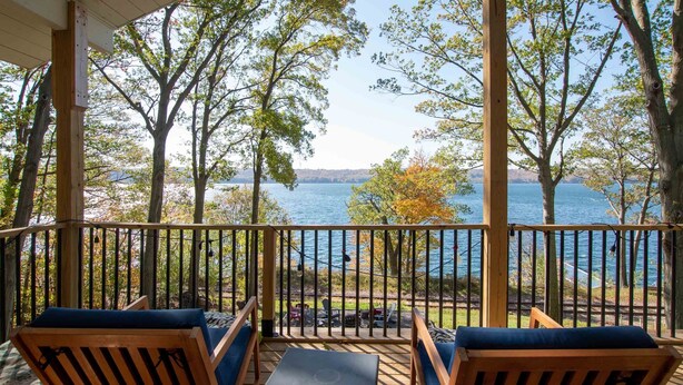 A covered deck with wooden railings, two wooden chairs with cushions, and a view of a lake with trees and a distant shoreline.