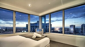 A modern hotel room with a large bed, a bedside table, and a view of the cityscape through large windows. - Holidays Gold Coast (Surfers Paradise)