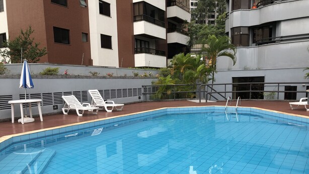 A rectangular swimming pool with a tiled edge, surrounded by a wooden deck. There are white lounge chairs and a blue and white umbrella. - Cozy and Cozy Apartment Portal do Morumbi (Vila Suzana)