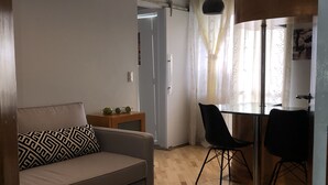 A living room with a sofa, a dining table with chairs, a hanging light fixture, and a door leading to another room. - Cozy and Cozy Apartment Portal do Morumbi (Vila Suzana)