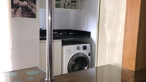 A compact kitchen with a washing machine, a stove, and a coffee maker visible through an open door. - Cozy and Cozy Apartment Portal do Morumbi (Vila Suzana)