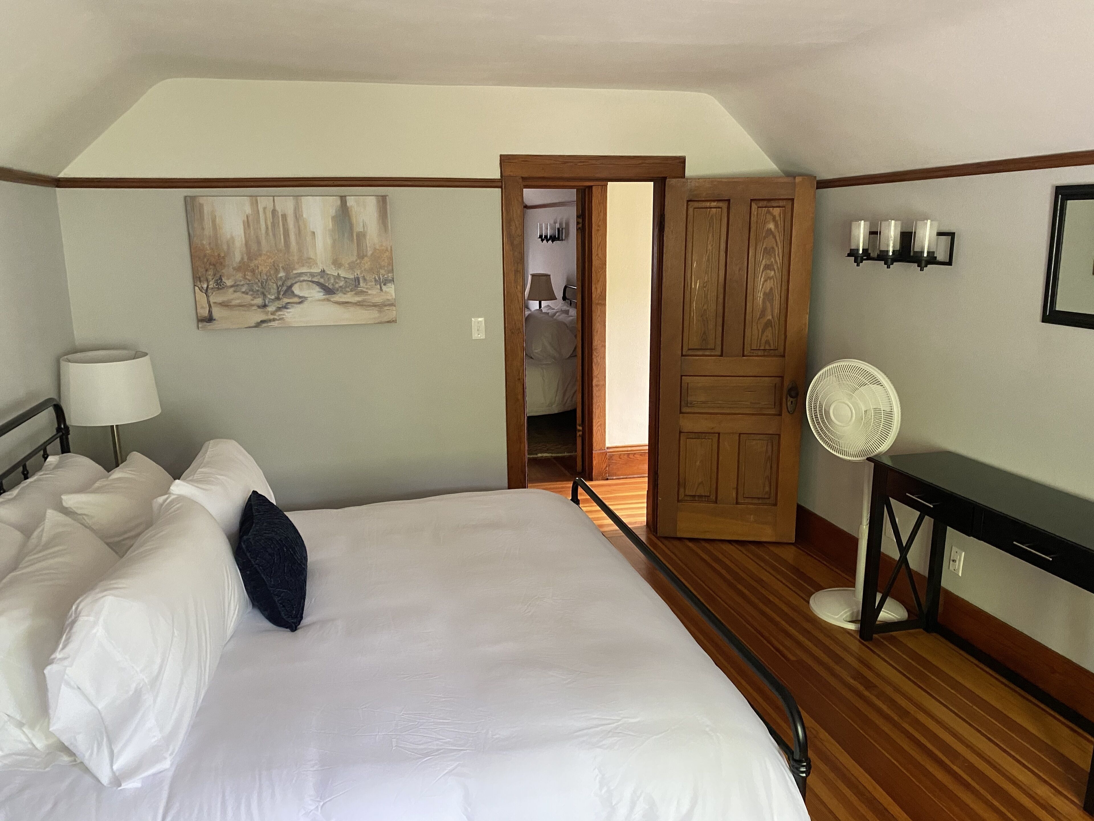 A hotel room with a large bed, a bedside table with a lamp, a painting on the wall, a wooden door, and a fan.