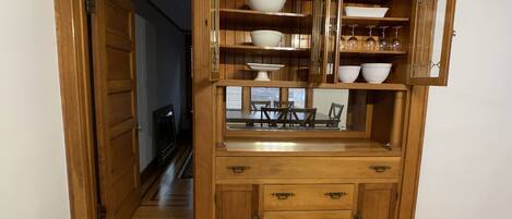 A wooden cabinet with multiple shelves and drawers, containing various kitchenware and a small dining table setup.