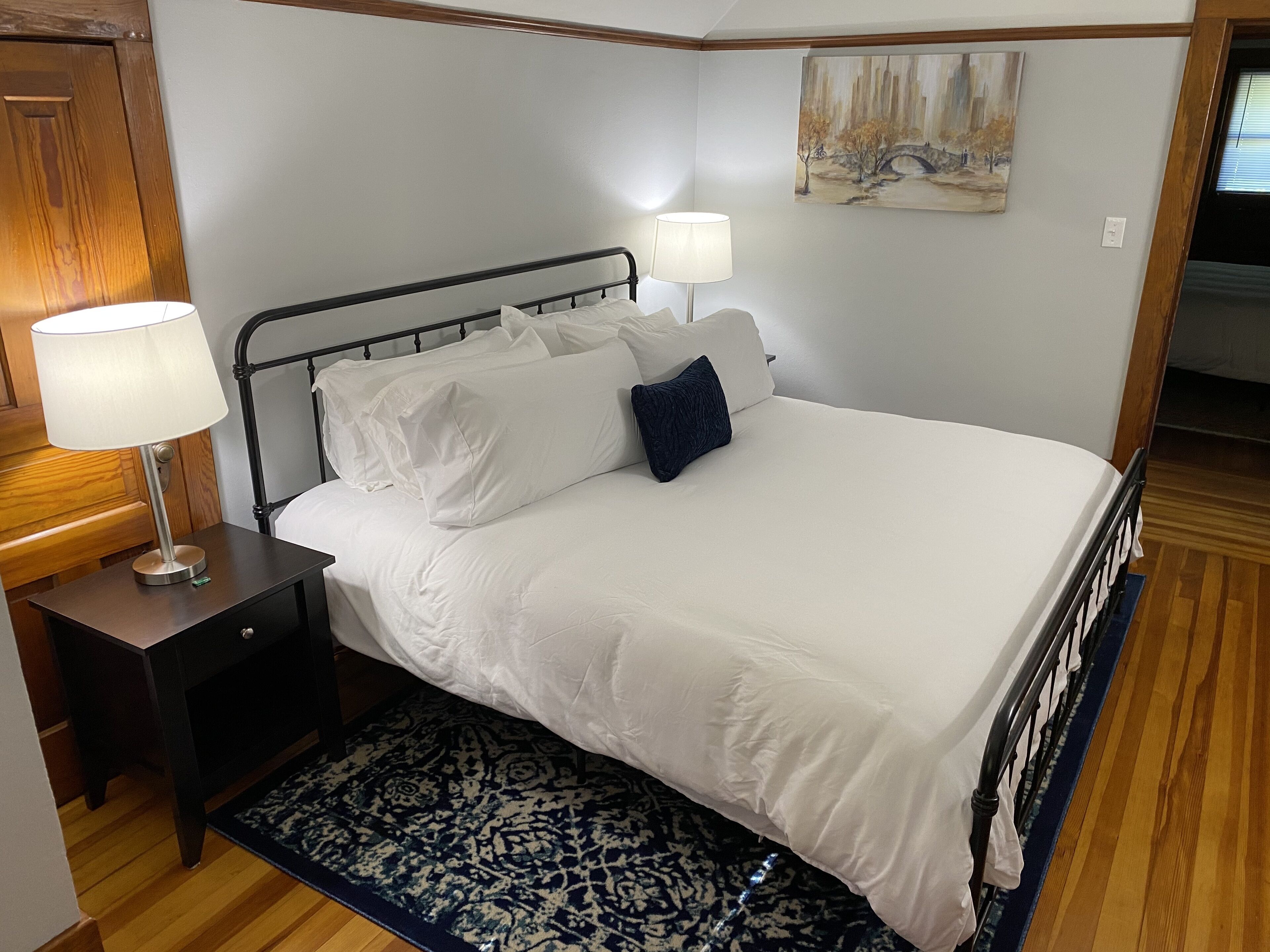 A neatly made bed with white linens and a blue throw pillow, a bedside table with a lamp, and a painting on the wall.