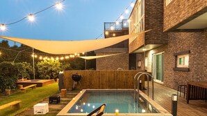 Un patio trasero con piscina, terraza de madera y luces colgantes.