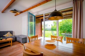 Un coin repas avec une table et des chaises en bois, un canapé agrémenté d’un coussin à motifs et une vue sur un jardin grâce à de grandes fenêtres.