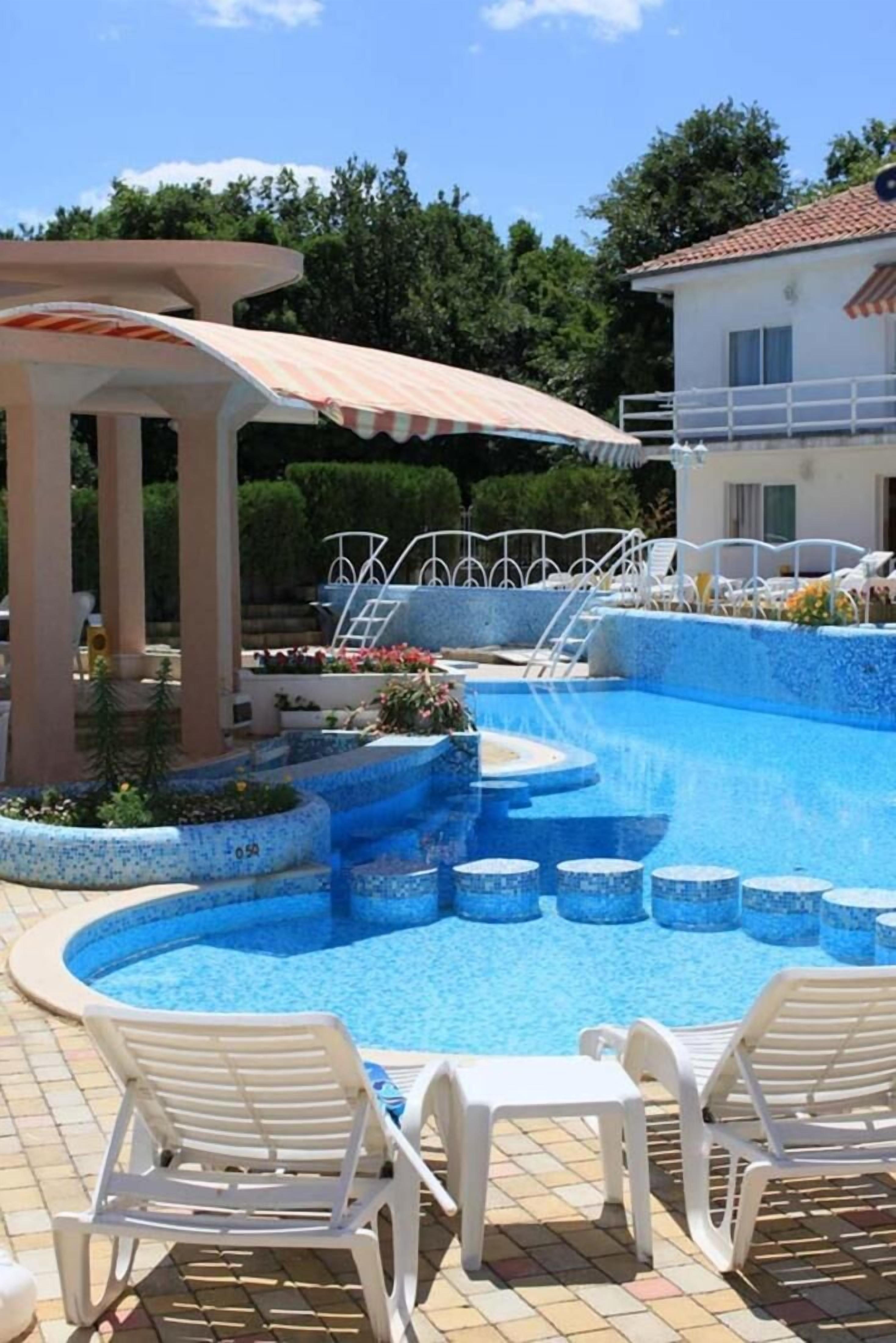 A swimming pool with a tiled deck, white lounge chairs, and a covered area with columns.
