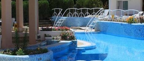 Una piscina con cubierta de baldosas, sillas de descanso blancas y una zona techada con columnas.