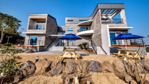 A modern multi-story building with a stone facade, multiple balconies, and outdoor seating areas with blue umbrellas. - Swimeong Spa Pension (Seogwipo)