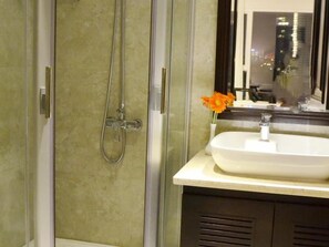 Une salle de bain moderne avec une douche à l’italienne en verre, un lavabo blanc et un miroir qui reflète le paysage urbain.