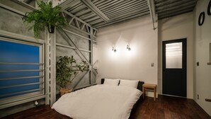 A bedroom with a large bed, a potted plant, a small table, and a door with a window. - Rakuna Inn Yu Kinshicho (Tokyo)