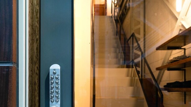 A door with a keypad lock, a metal handle, and a glass panel reflecting a staircase and interior lighting. - Rakuna Inn Yu Kinshicho (Tokyo)
