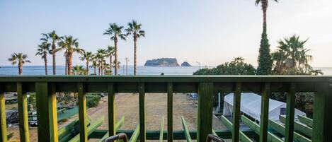 A wooden deck with a wicker chair, overlooking a beach with palm trees and a distant island.