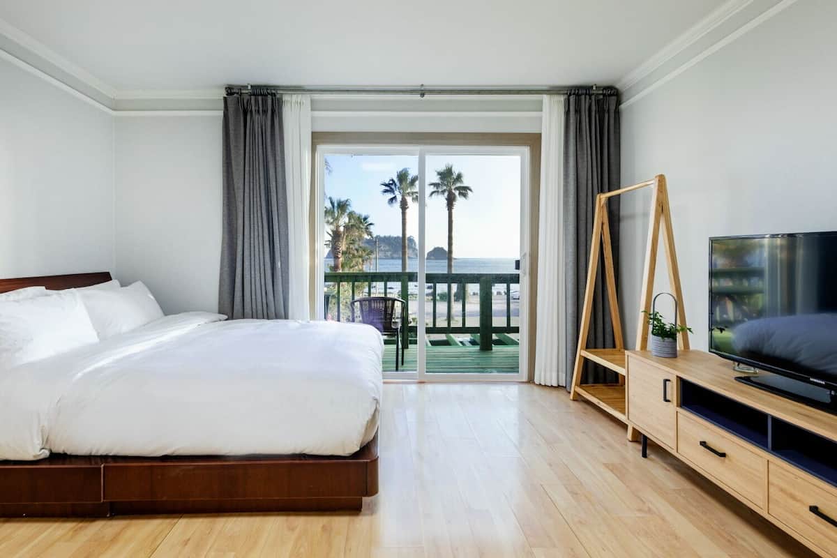 A hotel room with a bed, a TV, a wooden cabinet, and a balcony with a view of palm trees and the sea.