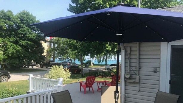 A covered patio with a glass table and chairs, a white railing, and a view of the street and trees.