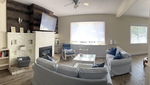 A living room with a fireplace, white brick fireplace, white sofa, blue armchair, and a flat-screen TV mounted on a wooden wall.