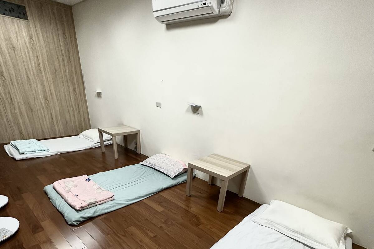 A room with two beds, a wooden floor, a wall-mounted air conditioner, and a small table.